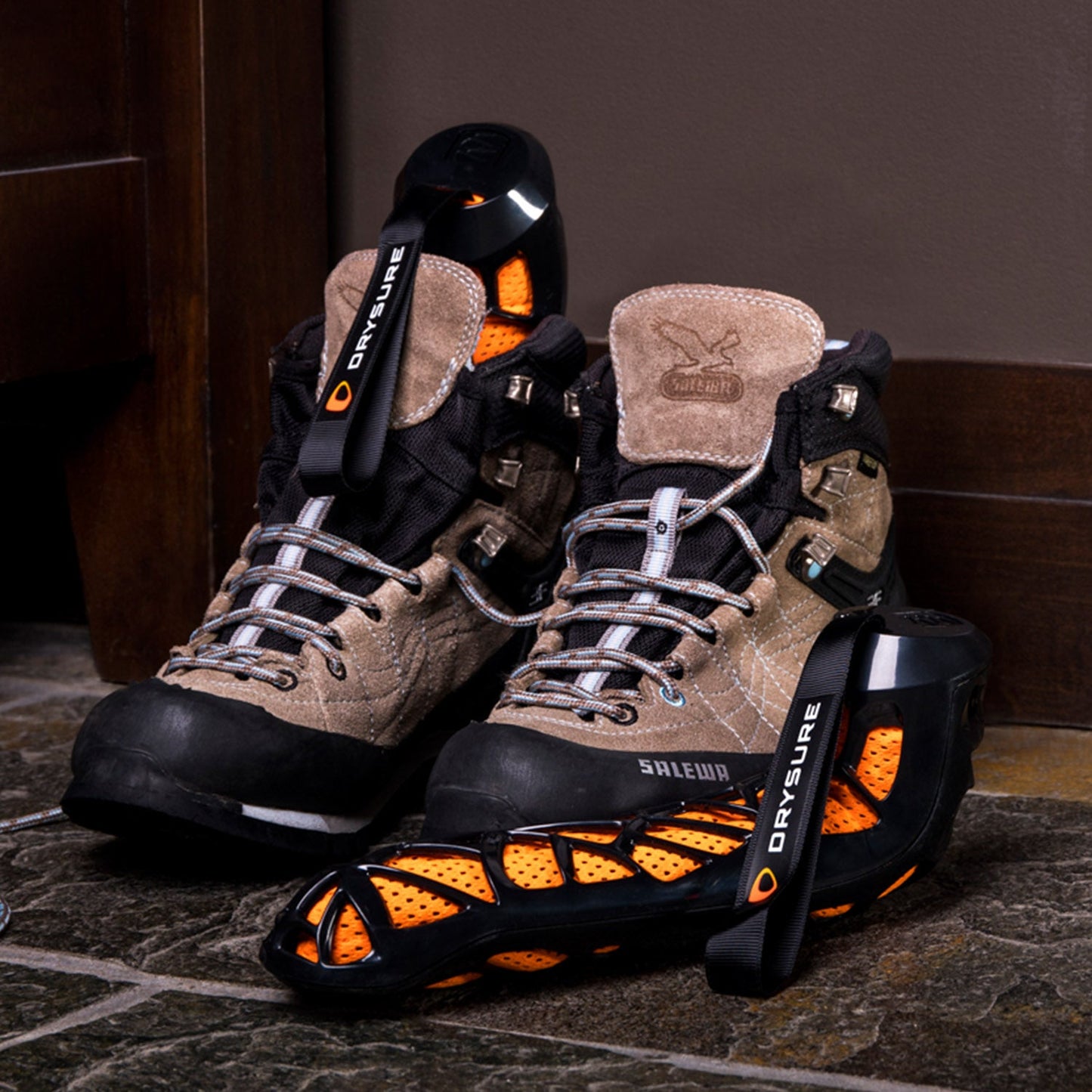 Pair of hiking boots with black and orange drysure Active boot dryers in one boot and in front of another on a stone floor.