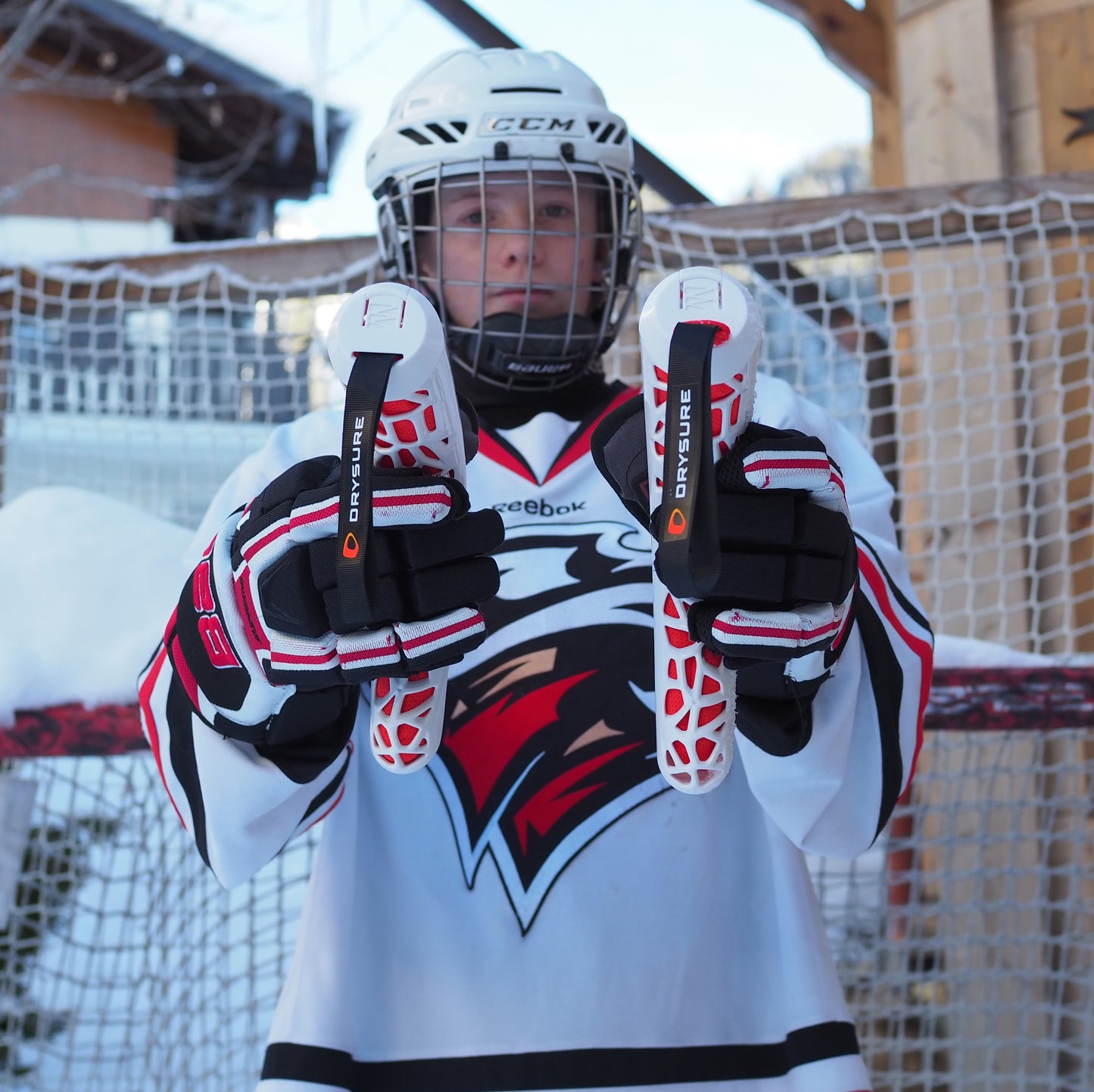 Person in hockey gear in front of a hockey net outdoors holding Drysure extreme boot dryers with visible brand logos in a snowy outdoor setting.